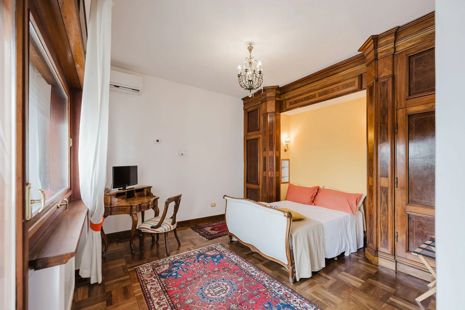 Camera con boiserie in noce, letto bianco con cuscini arancio e lampadario